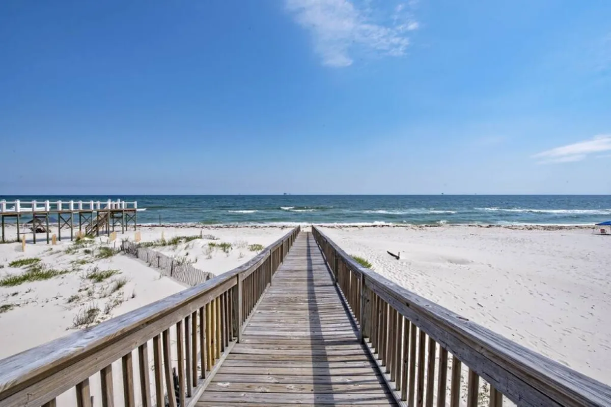 A picturesque wooden boardwalk leads from the house directly to the sandy beach, with the sparkling ocean visible ahead. The scene evokes a sense of anticipation and adventure, suggesting easy access for fishing trips, sunset strolls, or family gatherings by the shore.