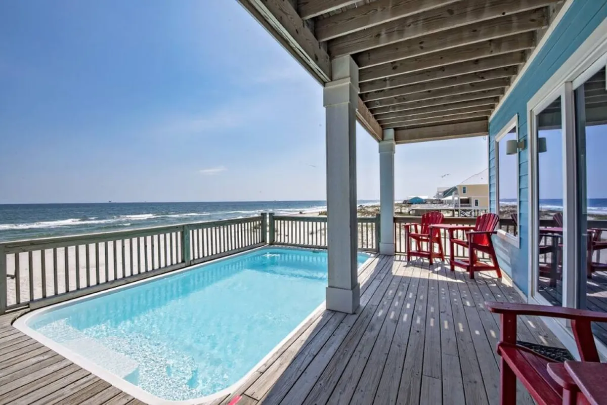 The image showcases a striking exterior scene with a private pool, ocean views, red deck chairs, and an expansive wooden deck. The scene radiates an atmosphere of vacation luxury, relaxation, and exclusive enjoyment with direct access to both pool and beach.