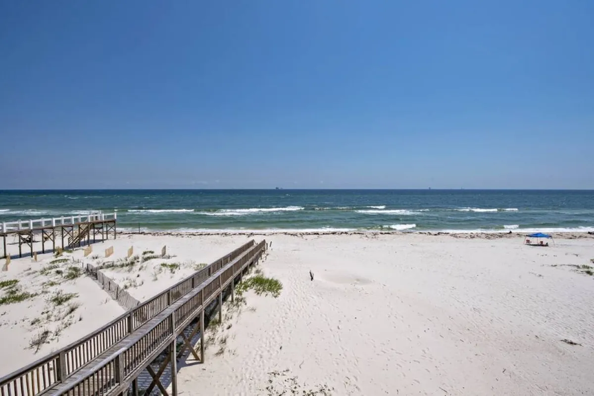 This serene exterior shot captures an expansive sandy beach, gentle ocean waves, and a boardwalk stretching toward the water. Distant ships on the horizon add to the sense of peaceful isolation.