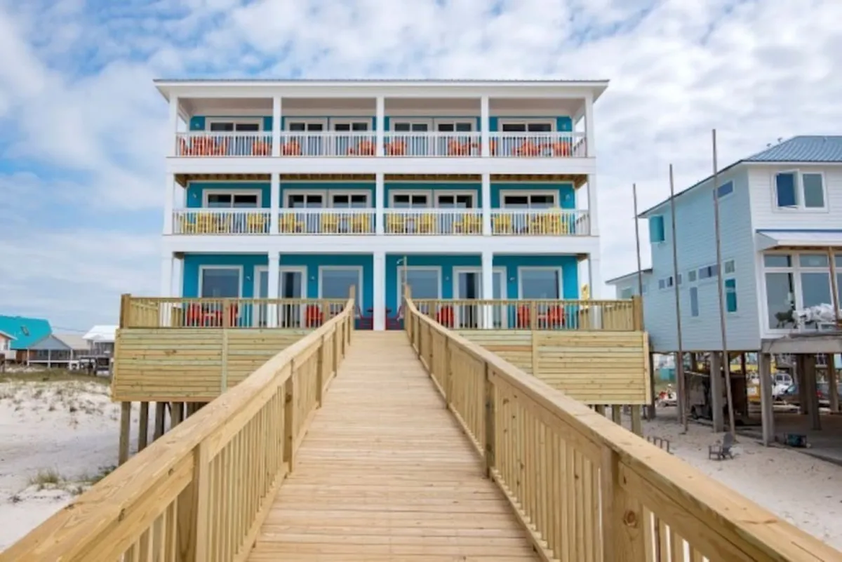 This image features the exterior of the property, displaying colorful balconies and a prominent wooden walkway leading directly to the beach. The large windows suggest open, airy interiors, while the scene captures the inviting atmosphere of the beachfront deck and private access to the sand.