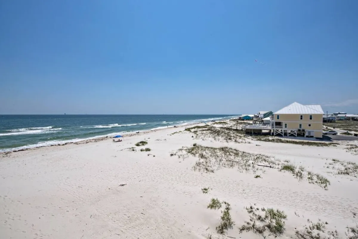 A wide view of an expansive, sandy beach with gentle ocean waves and a few distant houses dotting the background. The sun casts a warm light over the shore, creating an inviting atmosphere for a range of group activities such as beachcombing, swimming, or organizing a friendly beach Olympics.