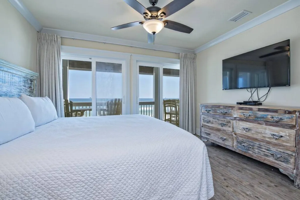 A serene interior featuring a king-size bed with large windows that frame an inviting ocean view, complemented by a rustic dresser and ceiling fan. The room exudes comfort and privacy, ideal for restful nights during a retreat.