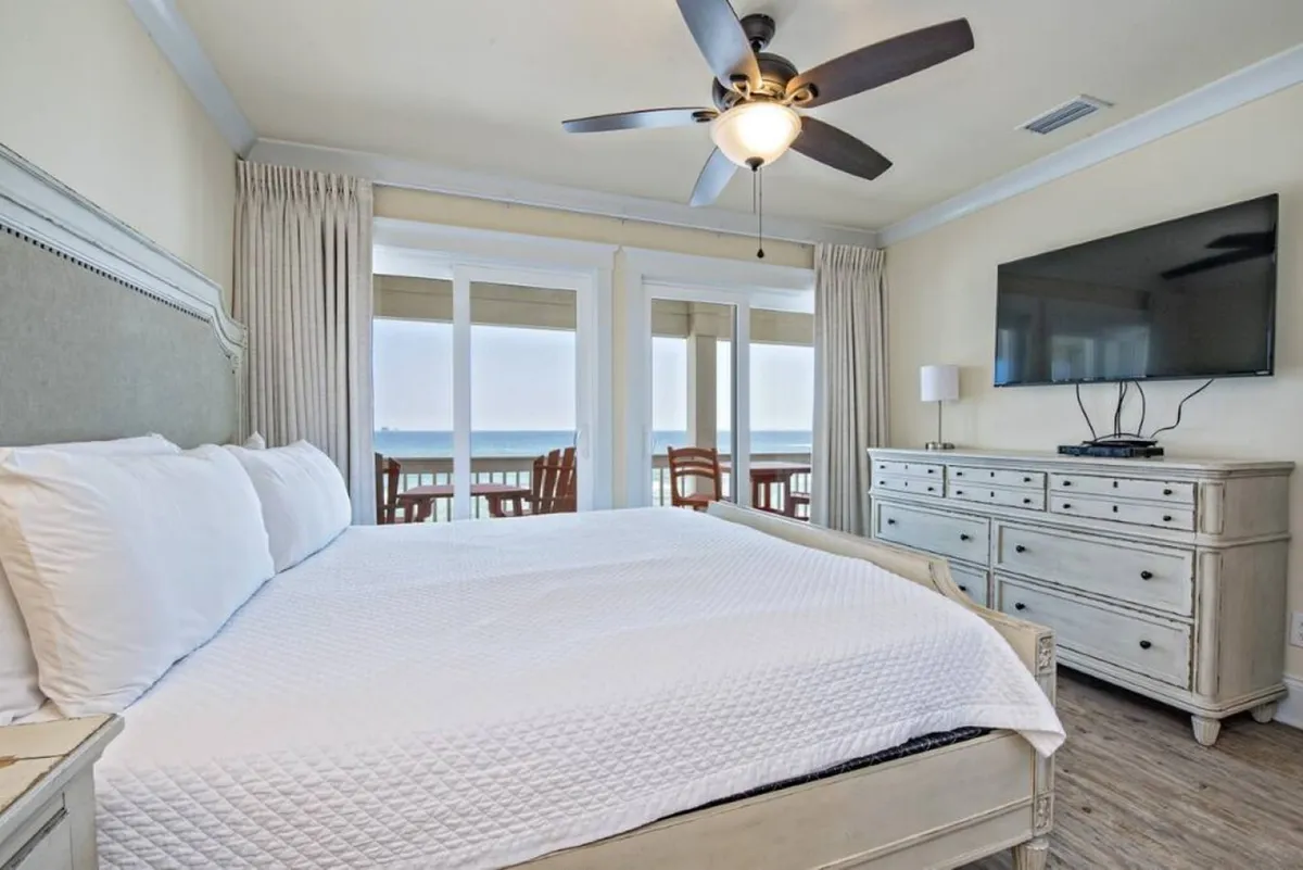 A spacious king bedroom with a king-sized bed facing large windows that offer an ocean view. The room is bathed in natural light and features a ceiling fan, TV, and dresser, conveying a restful and serene atmosphere.