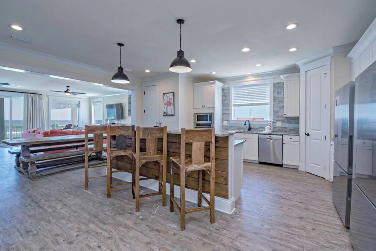 A bright, inviting kitchen and dining space with a kitchen island, wooden bar stools, a dining table, and large windows that offer ocean views. The setting is ideal for preparing meals, socializing, and enjoying the comforts of a modern, well-equipped home.