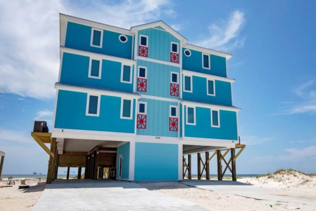 The hero image showcases the vibrant blue beachfront house raised on stilts, with expansive balconies and large windows overlooking the sugar-white sand and shimmering Gulf of Mexico. The relaxed, coastal scene features the private pool area, plenty of space for outdoor lounging, and a direct path to the tranquil, uncrowded shoreline stretching into the horizon.