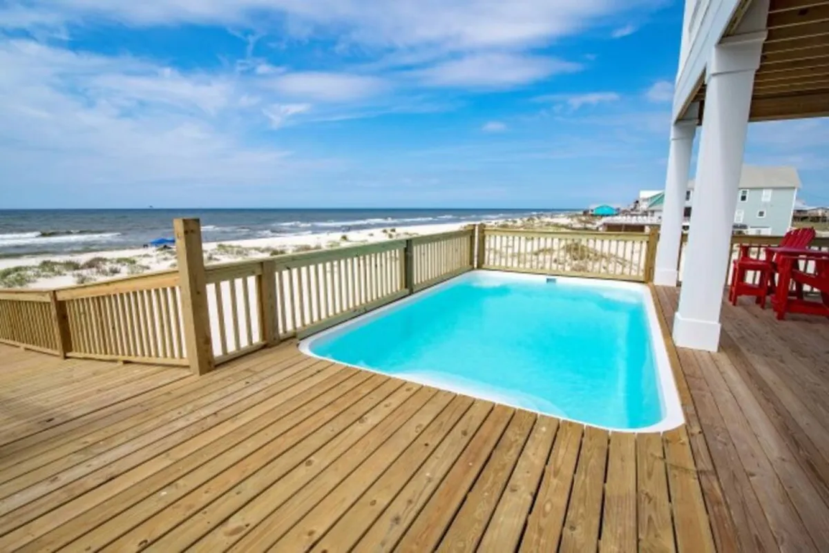 A private pool with clear blue water sits on a wooden deck overlooking the ocean. Bright red chairs add a pop of color and invite relaxation, while gentle waves and a wide sandy beach provide a stunning backdrop. The overall scene is peaceful and inviting, suggesting a blend of luxury and natural beauty.