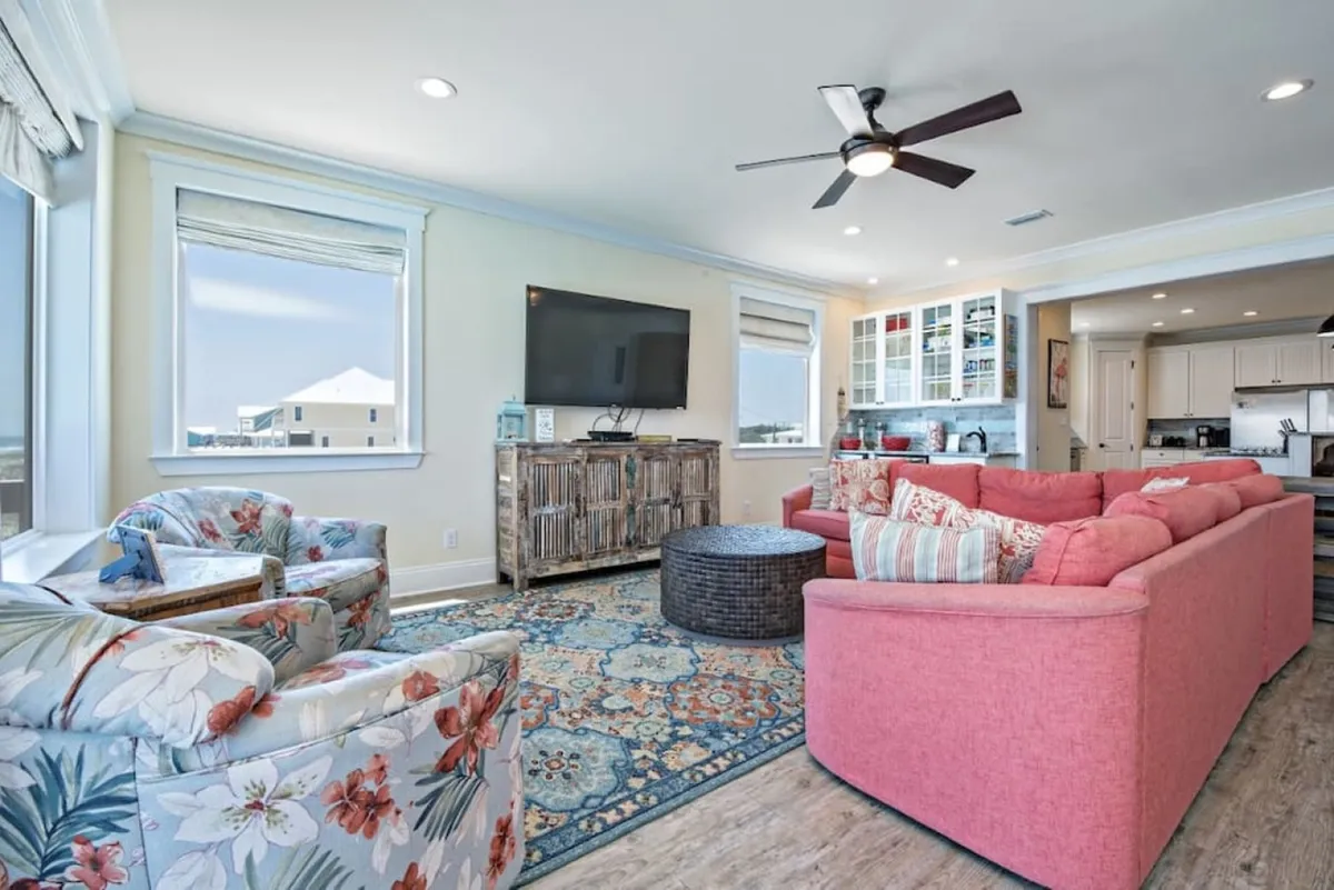 This vibrant living room features a large pink sectional sofa, floral armchairs, a flat-screen TV on a rustic wooden console, and a colorful area rug. The space is open and inviting, ideal for group relaxation or lively social events.