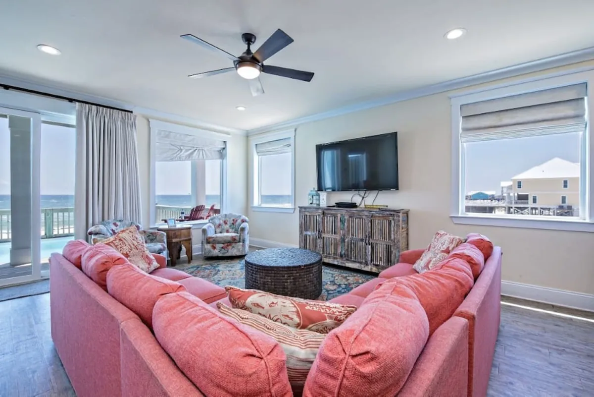 This interior shot presents a large coral-colored sectional sofa, a flat-screen TV, patterned armchairs, and sliding glass doors leading to a balcony with direct ocean views. The room is bright and welcoming, offering both relaxation and a visual connection to the outdoor decks and the Gulf beyond.
