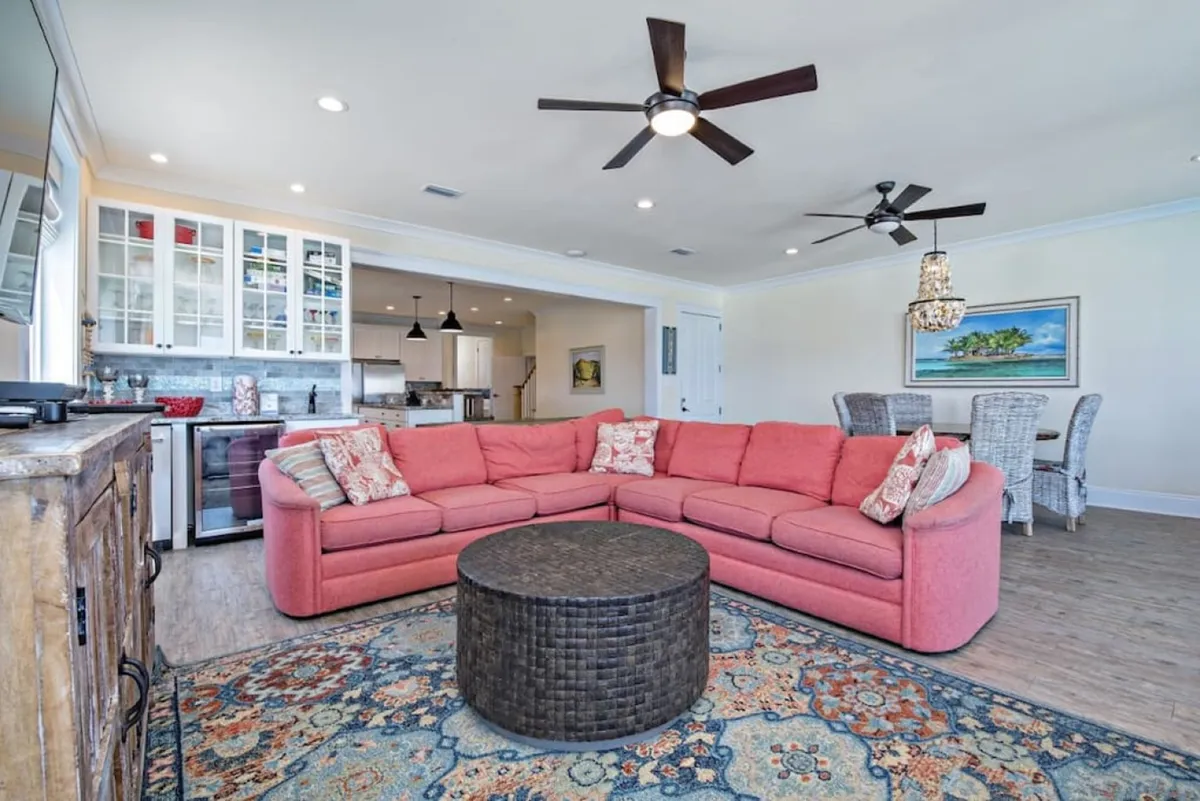 This image showcases a bright, open-concept living space featuring a large coral sectional sofa, a round woven coffee table, an open kitchen with white cabinetry, and a dining area with wicker chairs and a seashell chandelier. Large windows allow for abundant natural light and hint at ocean views beyond, reinforcing the airy, communal feel.