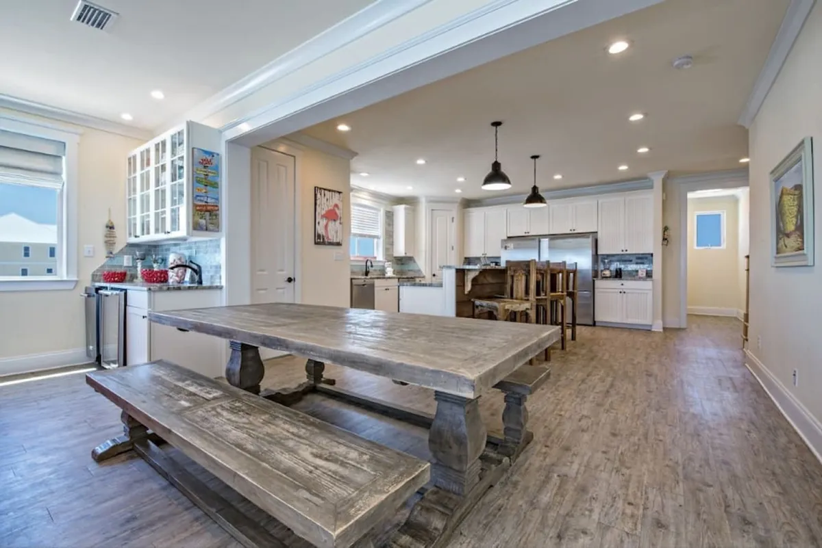 The scene features a rustic wooden dining table with benches, surrounded by modern white kitchen cabinetry, stainless steel appliances, and a kitchen island with wooden bar stools. The space feels warm and inviting, ideal for large family meals or group gatherings.