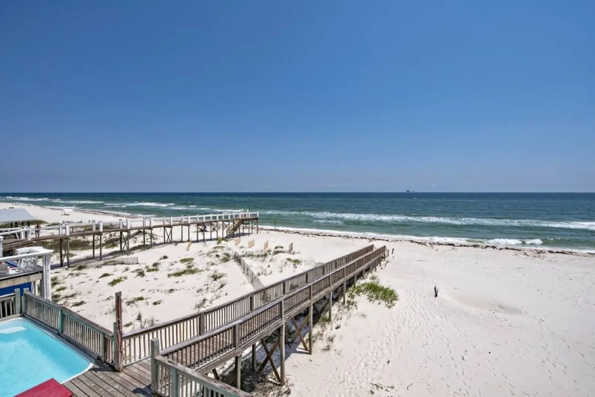 A tranquil scene featuring a private beachfront pool in the lower left, a wooden boardwalk leading directly to a secluded stretch of white sandy beach, and gentle blue ocean waves meeting the horizon. The scene evokes luxury and privacy, with ample space for group activities and relaxation right at the doorstep.