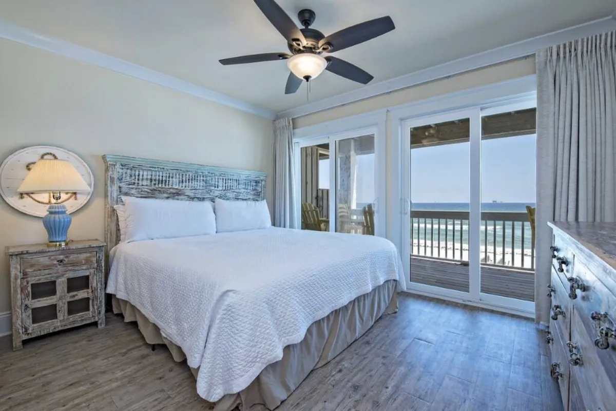 A tranquil king bedroom with white bedding, rustic wooden furniture, and large sliding glass doors opening to a balcony with panoramic ocean views. A decorative table lamp with a blue base adds a coastal touch.