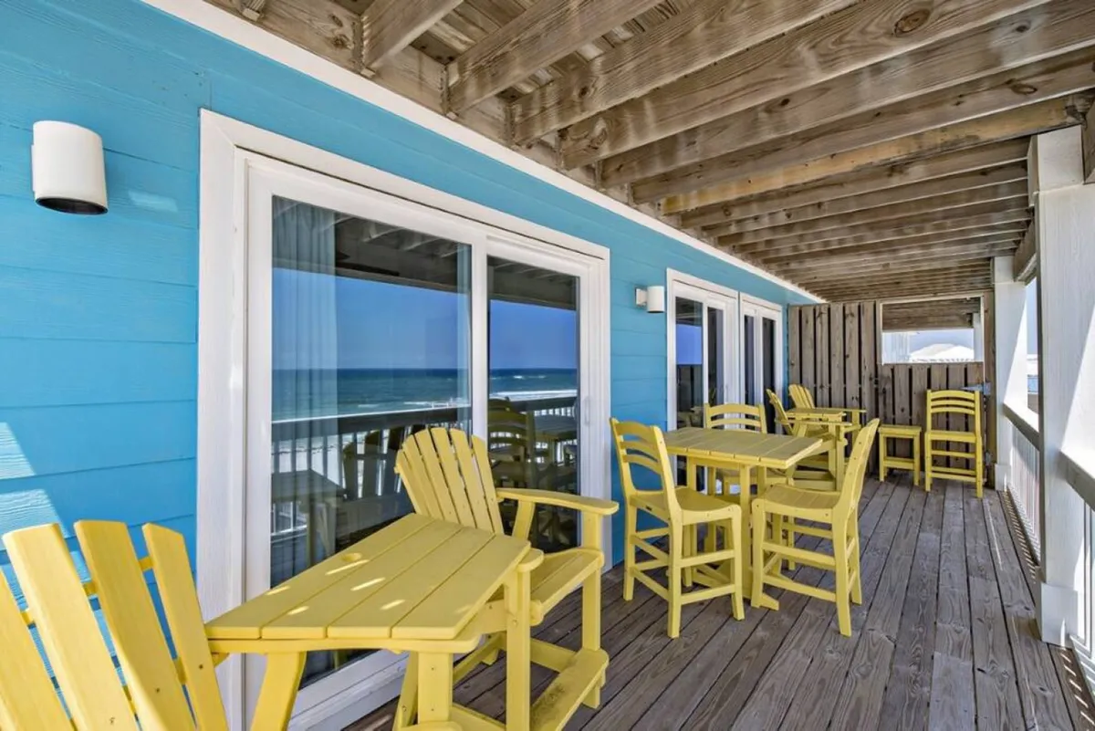 A spacious oceanfront balcony featuring yellow Adirondack and dining chairs, a blue painted exterior wall, and wooden deck flooring. The ocean view is reflected in sliding glass doors, reinforcing the connection between indoor and outdoor living spaces.