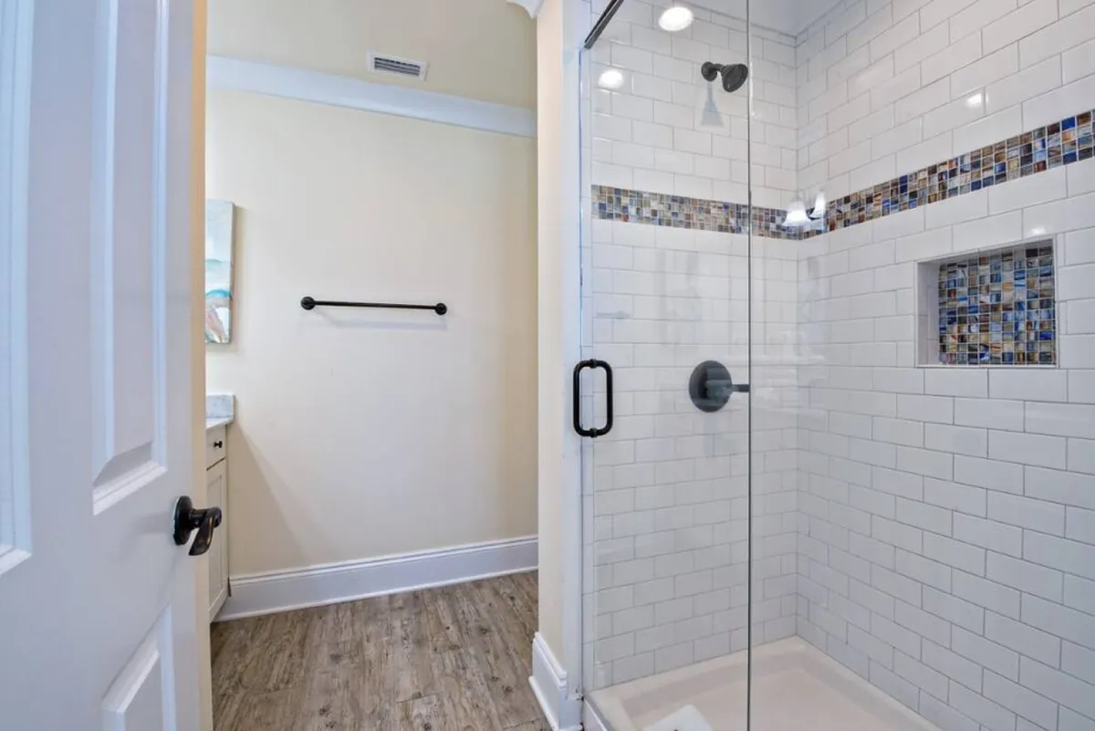 A modern bathroom with a glass-enclosed shower adorned with tile mosaic accents, sleek white subway tiles, wood-look flooring, and a stylish vanity visible in the background. The space is bright, fresh, and meticulously maintained.