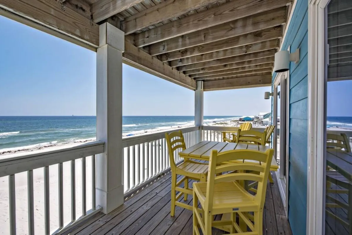 A wide balcony with yellow chairs and a wooden table, exposed beams overhead, and an expansive view of the sandy coastline and rolling waves. The open layout invites relaxation and socializing, with the ocean stretching out just beyond the railing.