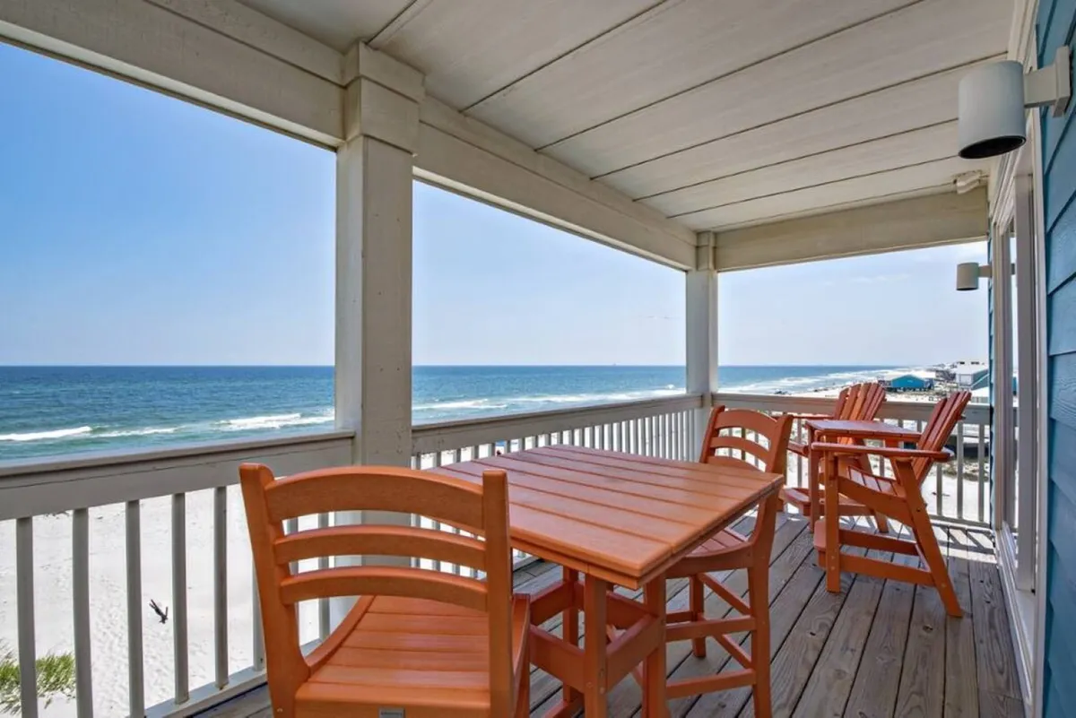 A serene balcony or deck area with a wooden dining table and chairs, framed by a white railing and offering a stunning direct view of the ocean. The space is set up for outdoor dining and relaxation, perfect for both sunrise breakfasts and sunset gatherings.