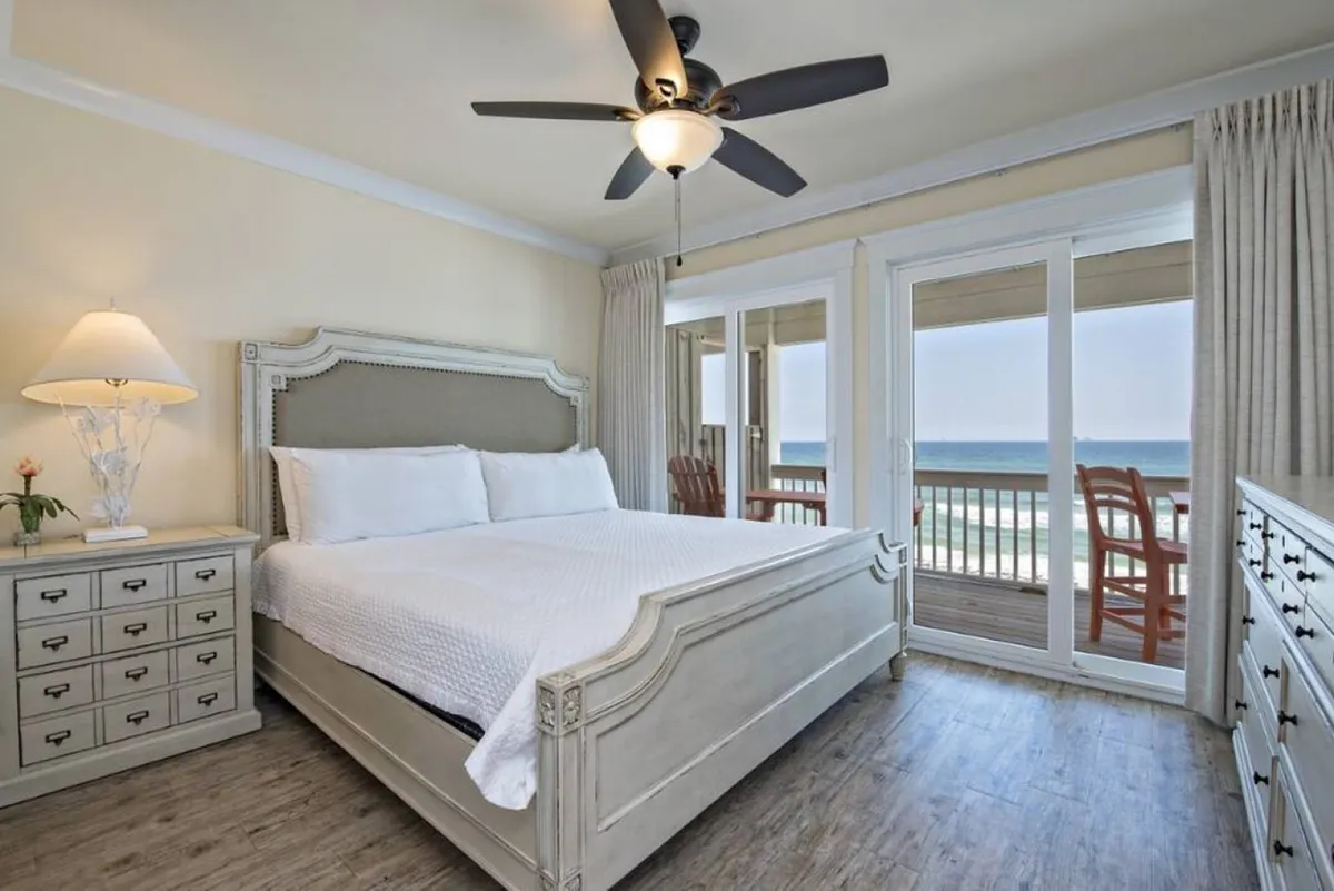 A spacious king-sized bedroom with a textured headboard, floor-to-ceiling glass doors leading to a private balcony, and wooden chairs set for enjoying panoramic ocean views. The room is elegantly furnished and bathed in natural light, creating a peaceful sanctuary.