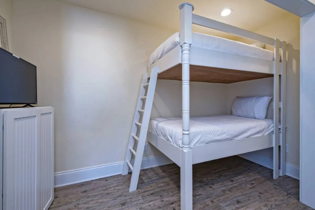 A cheerful and comfortable bunk room featuring a white wooden bunk bed, a small TV on a cabinet, wooden flooring, and soft wall colors. The space is practical and inviting, ideal for children or younger guests seeking their own retreat.