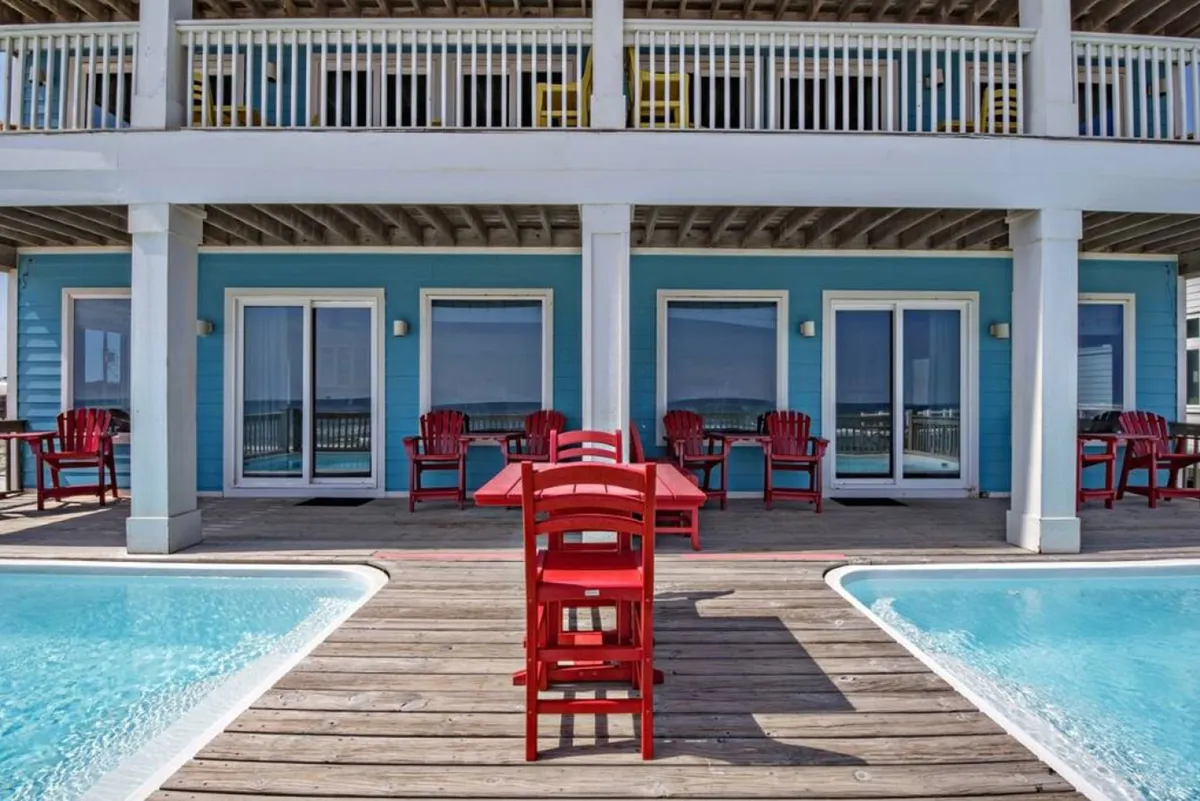 A vibrant, cheerful scene featuring a private pool with turquoise water, bright red outdoor chairs and table, the blue exterior walls of the house, and a wooden deck area. The setting is lively and inviting, ideal for group socializing and relaxation with the ocean close by.