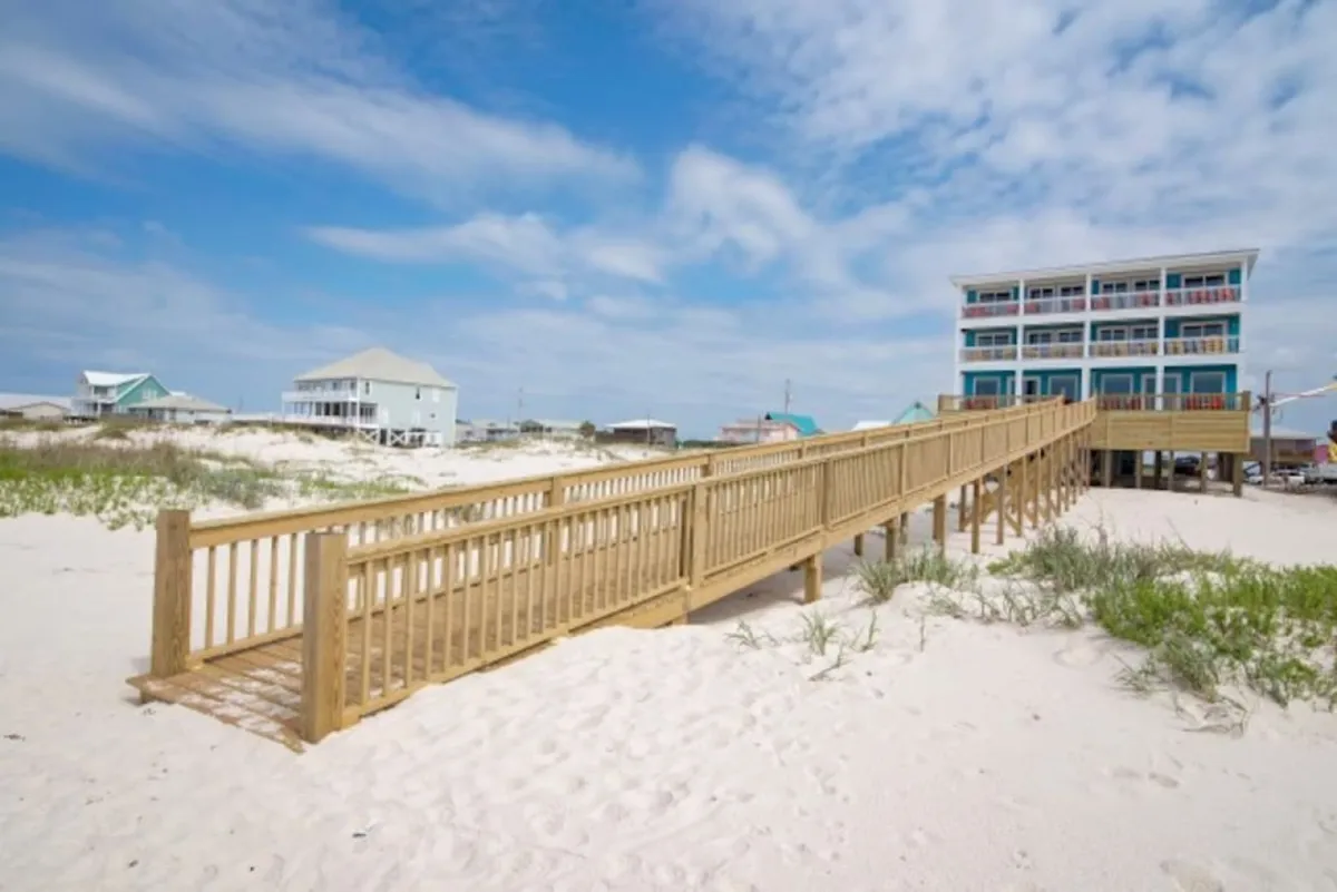 A wide view featuring a wooden boardwalk leading to a large multi-story beach house, set against an expansive stretch of white sandy beach. Distant beach houses can be seen in the background, emphasizing both the privacy and scale of the property, while the scene evokes a sense of tranquility and exclusivity.