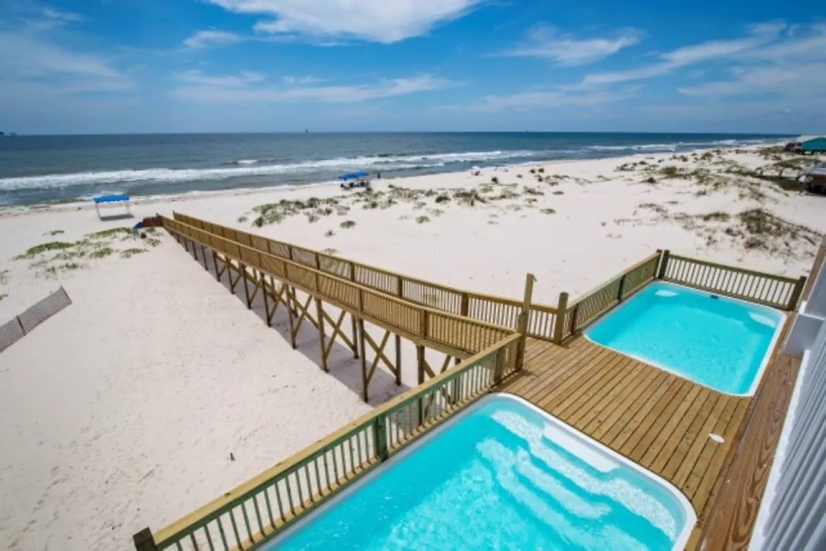 Private beachfront swimming pools sit in the foreground, with a wooden boardwalk leading past them directly to an expansive sandy beach and the Gulf's blue horizon. The scene is peaceful, with sunbathing areas and ample space for outdoor relaxation or group activities.