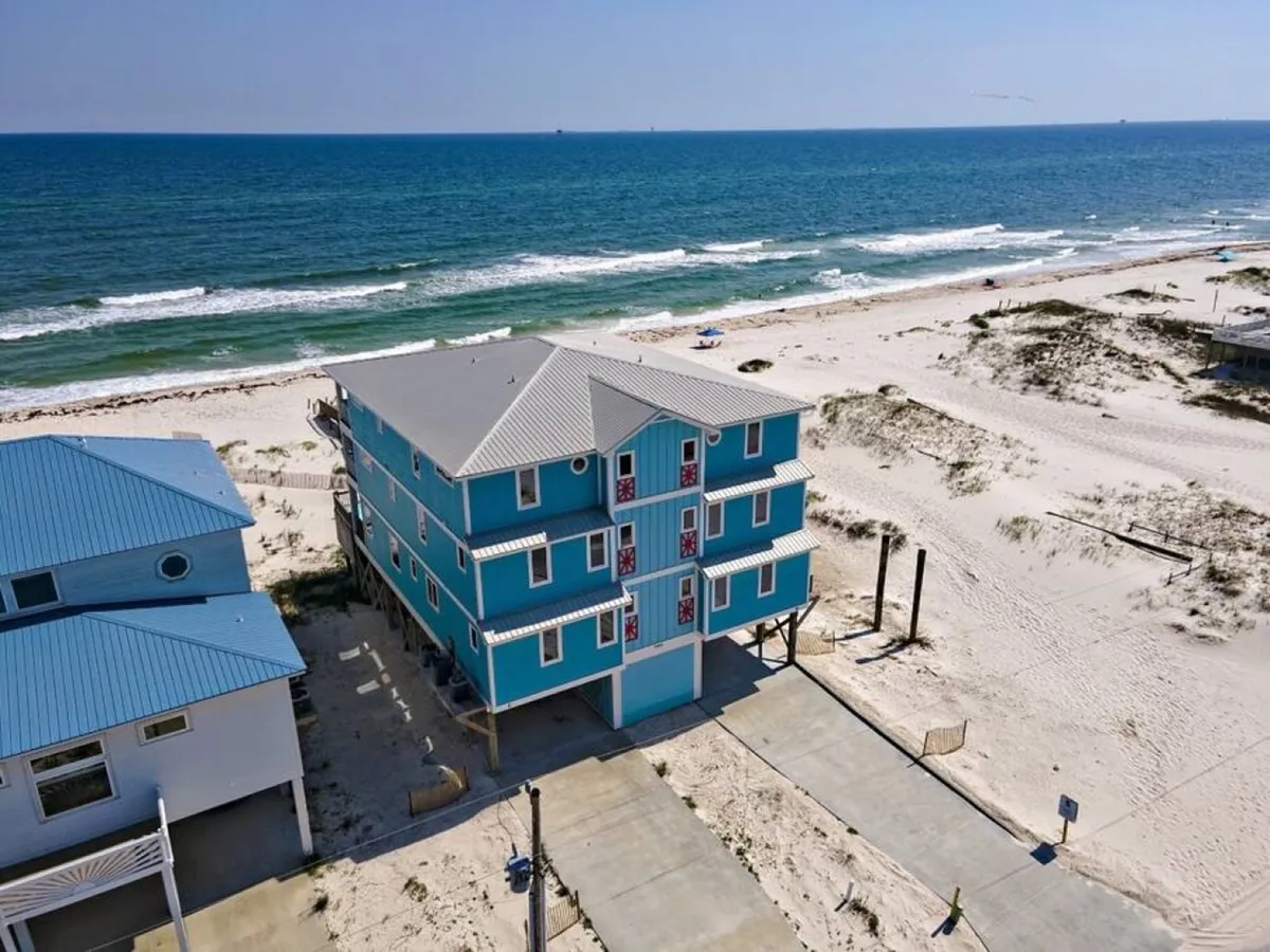 A panoramic exterior of a luxurious, bright blue three-story beachfront house, set directly on an expansive, pristine white beach. The calm ocean stretches into the horizon and neighboring homes are visible, but the focus is on the seclusion and grandeur of the property.