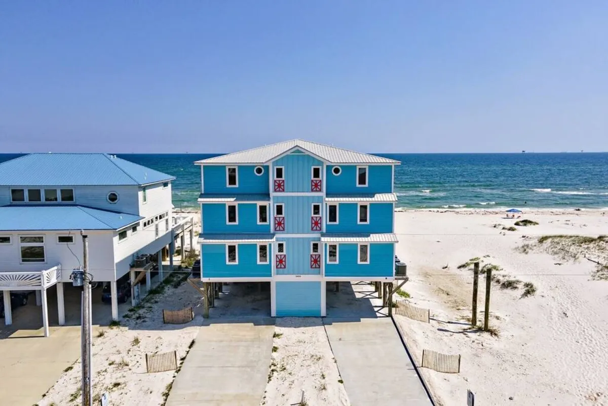This exterior image shows the bright blue beachfront house with red accents, a wide concrete driveway for ample parking, and a private sandy beach leading to the Gulf. The house’s multiple balconies are visible, suggesting spacious accommodations for large groups or events.