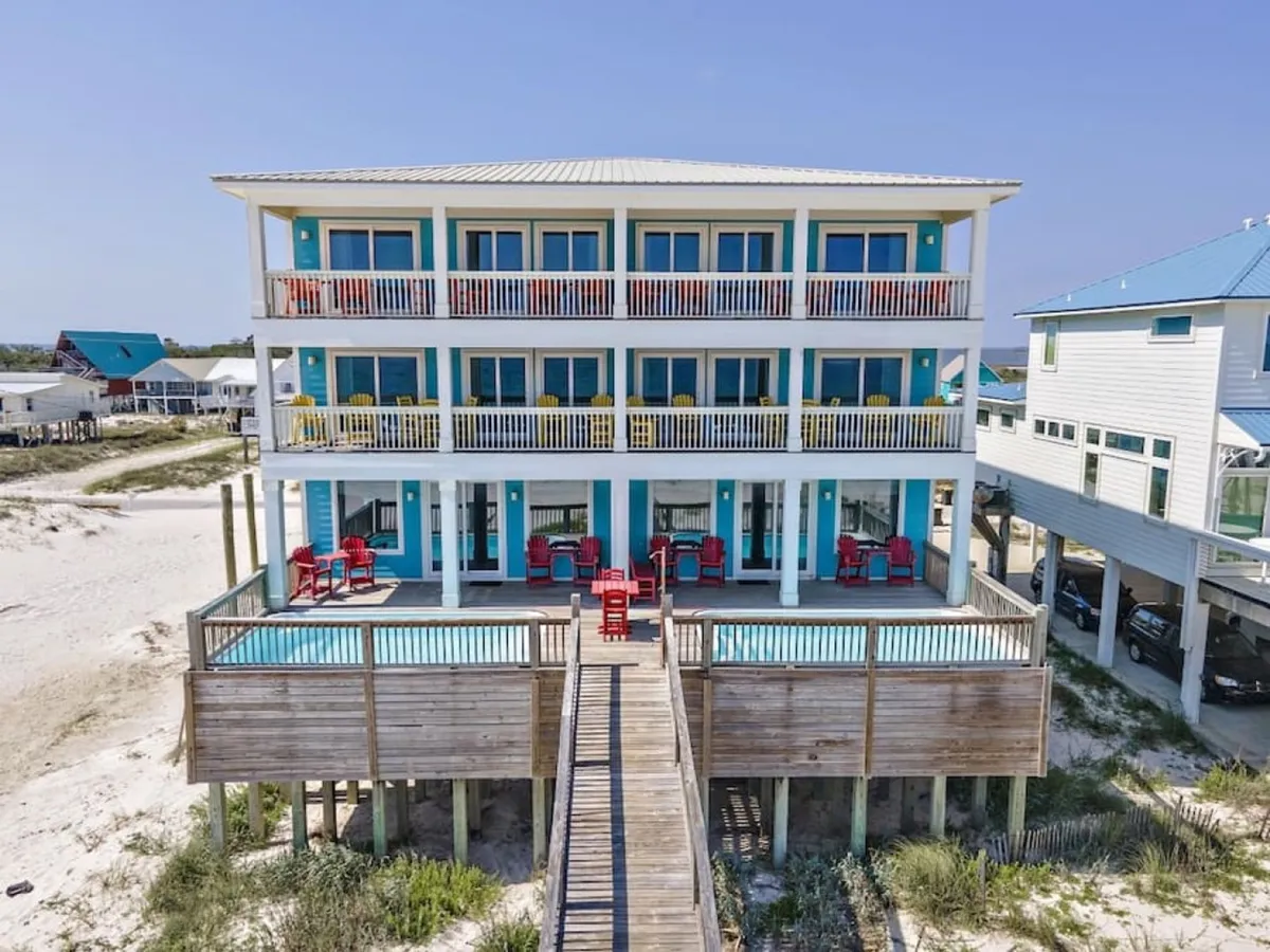 A vibrant three-story beachfront house with spacious balconies facing the water, a private pool on the ground-level deck, and red Adirondack chairs arranged for relaxation. A wooden walkway stretches from the house directly to a wide, sandy beach, emphasizing the direct access and private nature of the location.