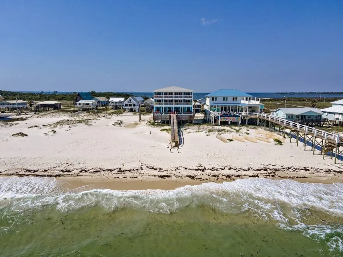A large beachfront duplex with multiple balconies overlooks a sandy beach and gentle ocean waves. The property has a private walkway leading directly to the beach, and neighboring beachfront houses hint at a peaceful, exclusive neighborhood. The scene exudes tranquility and showcases the property's capacity for large groups and events.
