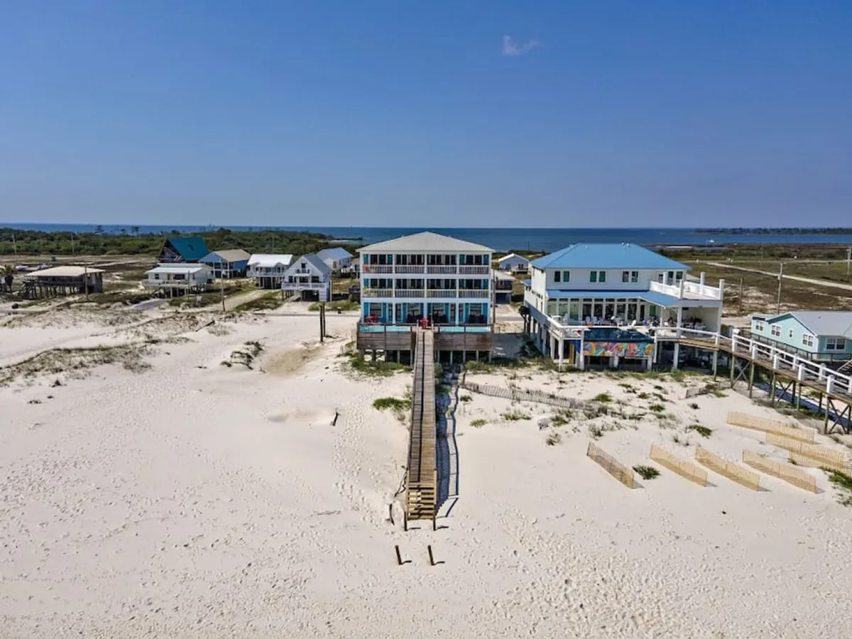 A striking view of a three-story beachfront house with spacious balconies, a long wooden walkway stretching to an expansive sandy beach, and the Gulf of Mexico in the background. The property is set apart from neighboring homes, highlighting a sense of privacy and luxury ideal for large groups.
