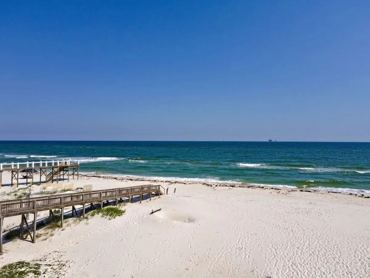 A pristine boardwalk leads directly to a white sand beach and calm blue ocean. The expansive sky and gentle waves create a peaceful, rejuvenating atmosphere, while the absence of crowds amplifies the sense of seclusion and tranquility.