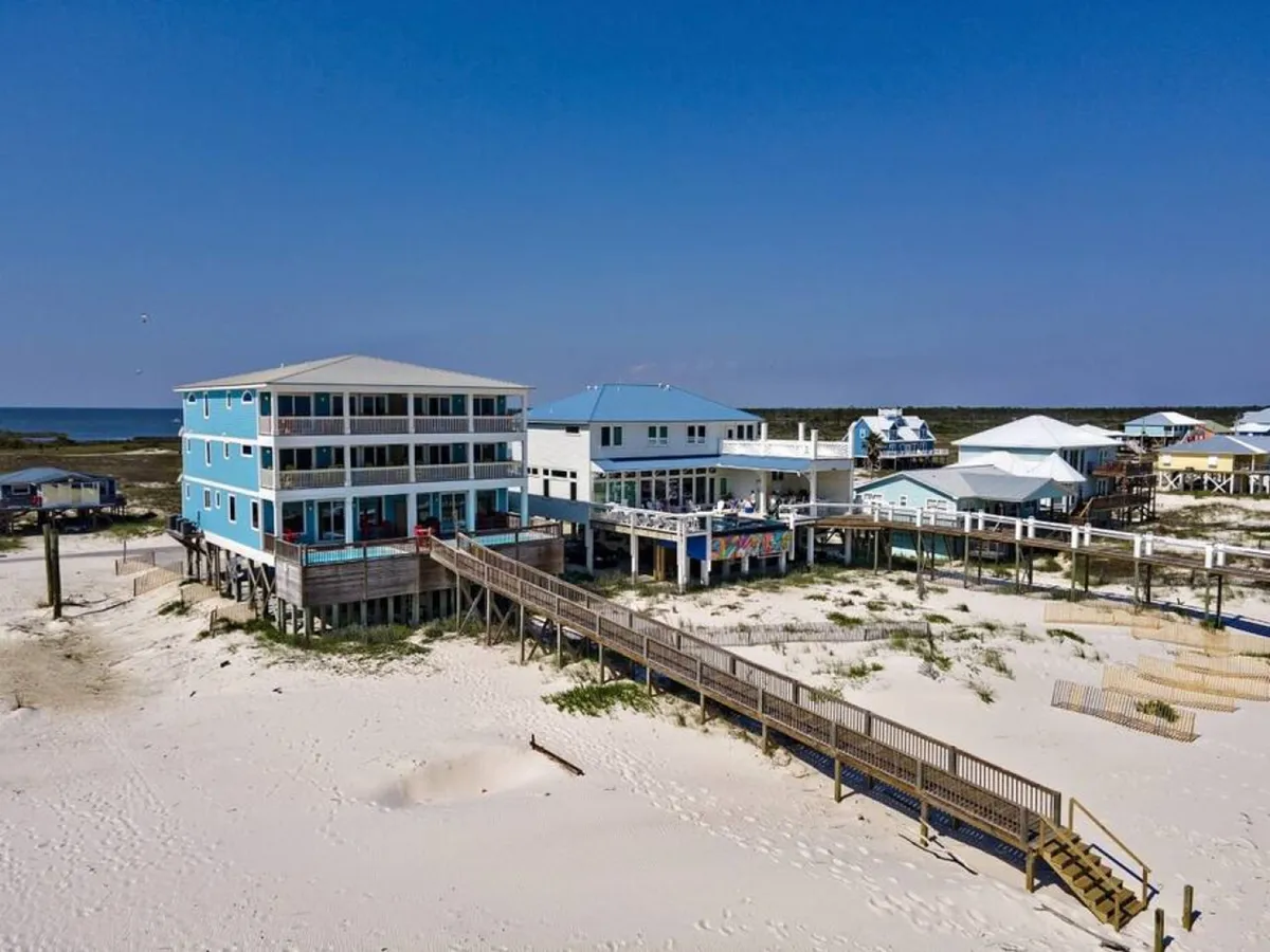 A three-story beachfront duplex with spacious balconies is framed by a wooden boardwalk leading onto a quiet sandy beach. The adjacent beach houses contribute to a sense of peaceful community, while the architecture hints at ample space and luxury for group stays.