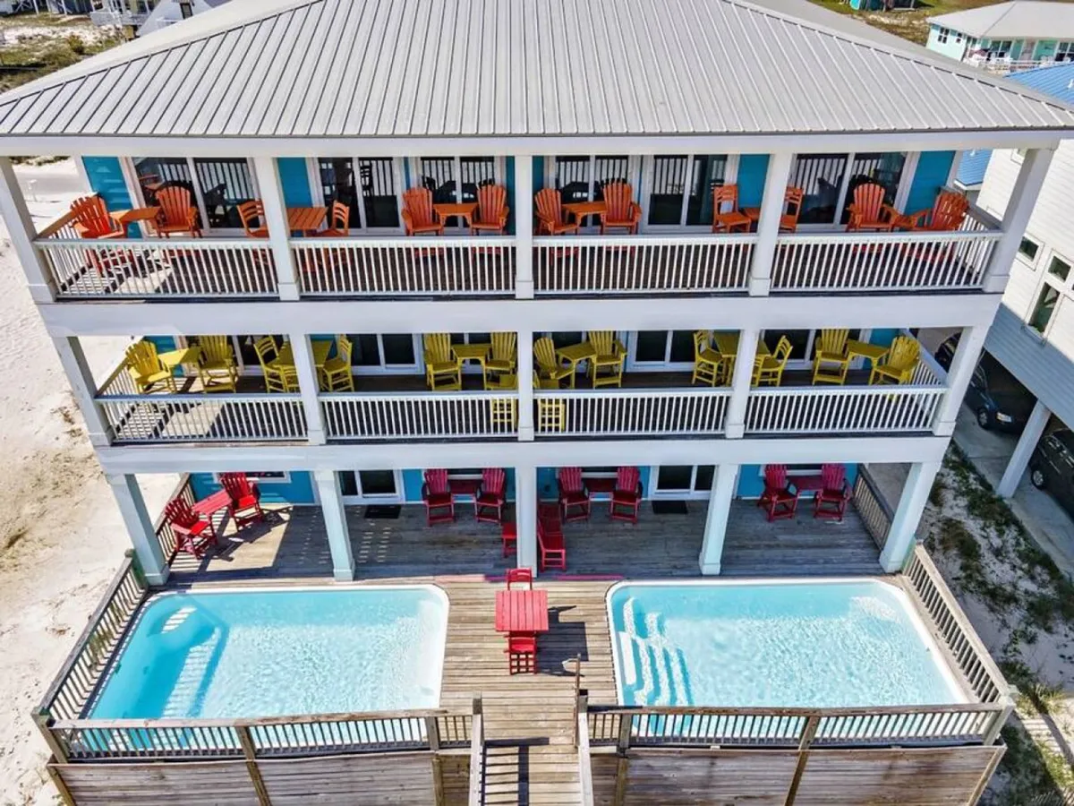 Three levels of balconies adorned with colorful Adirondack chairs overlooking two private swimming pools and extensive wooden deck areas, all set against the backdrop of the white sandy beach and Gulf waters.